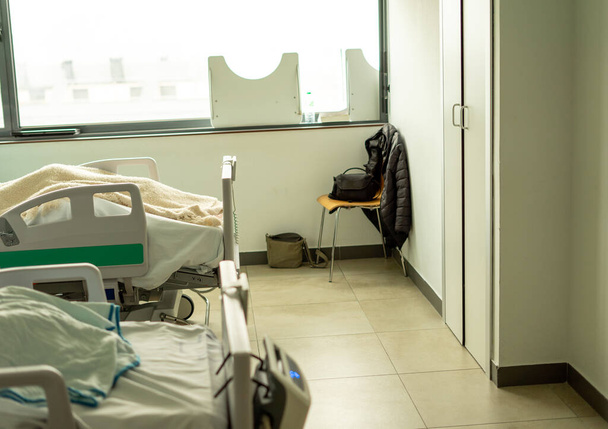 больничная палата с двумя кроватями, занятыми пациентами и на заднем кресле с сопровождающей одеждой - Фото, изображение