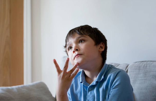 Portret van een jonge jongen die diep in gedachten uitkijkt met de hand op de kin, kind dat alleen zit met een denkend gezicht, kind dat tv kijkt of thuis ontspant na school - Foto, afbeelding