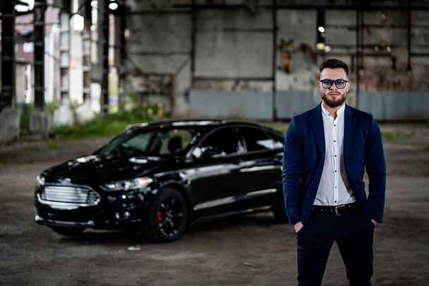 Stylish handsome man with sunglasses in fashionable clothes standing with the luxury black car on the background. Closeup - Фото, зображення