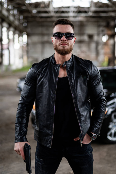 Man with a beard in a black leather jacket, wearing sunglasses and holding a gun. Man is looking at the camera. Guns concept - Foto, Imagen