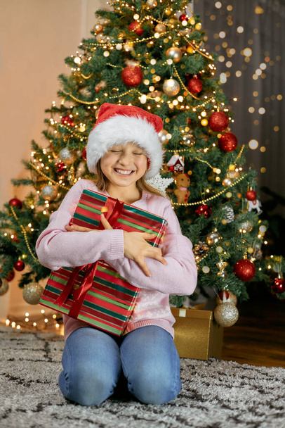 Happy New Year and Merry Christmas. A cute girl is very happy with the gift, hugs it and smiles with happiness - Photo, Image