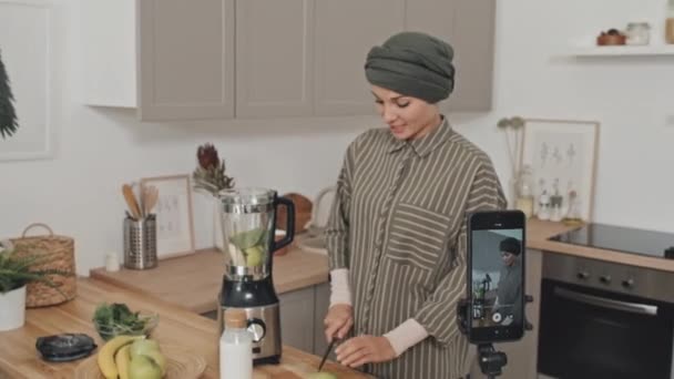 Moslim vrouwelijke blogger in hijab snijden van fruit en praten op camera tijdens het filmen van video recept met smartphone in de keuken - Video