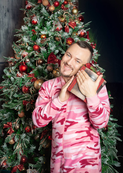 Cheerful funny happy man in pink sleepwear with Christmas present box standing near decorated fir tre - Photo, Image