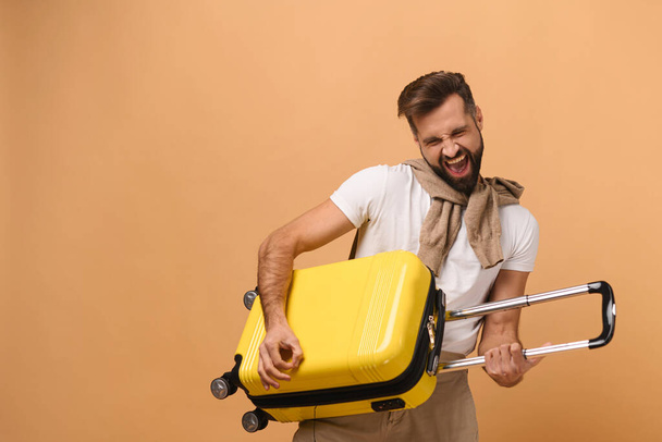 Cheerful caucasian man standing with yellow suitcase, playing on a bag like a guitarist, fooling around, while looking forward a trip isolated on orange - Фото, зображення
