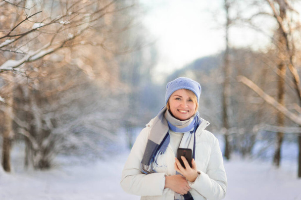Portrait of attractive woman in winter clothes, a blue hat and a white coat with phone in her hands. A woman uses her phone during a winter walk in a snow-covered park on a frosty sunny winter day. - Foto, Bild