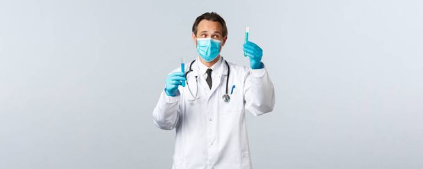 Covid-19 、ウイルス、医療従事者および予防接種の概念を防止する。臨床検査室の思慮深い医師は、医療用マスクと手袋を着用し、ワクチンサンプルを含む2つの検査管を見てください。 - 写真・画像