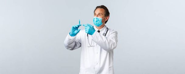 Covid-19 、ウイルス、医療従事者および予防接種の概念を防止する。医療用マスクと手袋を保持注射器で中年の医師は、コロナウイルスワクチンやHIV準備と、患者のために撮影準備 - 写真・画像