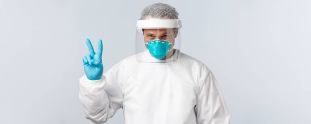 Covid-19, preventing virus, healthcare workers and vaccination concept. Determined doctor in personal protective equipment fighting coronavirus disease, show v-sign or peace, treating sick patients - Foto, Imagen