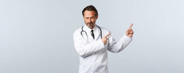 Covid-19, ξέσπασμα του ιού της στέψης, επαγγελματίες υγείας και πανδημία. Σκεπτικός και αβέβαιος άνδρας γιατρός, γιατρός με λευκό παλτό, συνοφρυωμένος αμφίβολος και δείχνοντας επάνω δεξιά γωνία - Φωτογραφία, εικόνα