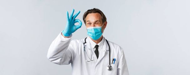 Covid-19 、ウイルス、医療従事者および予防接種の概念を防止する。医療マスクや手袋で陽気満足医師は大丈夫なサインとウィンクを表示します.医師は承認を与え、患者に同意する - 写真・画像