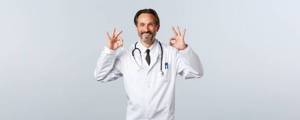 Covid-19, ξέσπασμα του ιού της στέψης, επαγγελματίες υγείας και πανδημία. Ικανοποιημένος χαρούμενος γιατρός εγγυάται την ποιότητα. Ψυχολόγος δείχνουν εντάξει χειρονομία και χαμογελώντας ευχαριστημένος, διαφημίζουν promo - Φωτογραφία, εικόνα