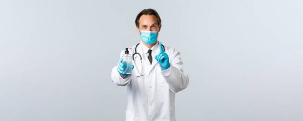 Covid-19, virüsü önleme, sağlık çalışanları ve aşı konsepti. Hevesli gülümseyen doktor el dezenfektanı kullanmanın önemini açıklar, birinci kuralı gösterir, tıbbi maske takar ve eldiven giyer. - Fotoğraf, Görsel