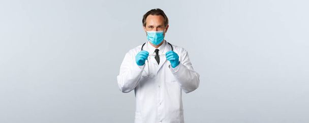 Ковид-19, профилактика вируса, медицинские работники и концепция вакцинации. Серьезный врач в медицинской маске и перчатках надевает очки, лечит больных пациентов, проверяет людей в клинике
 - Фото, изображение