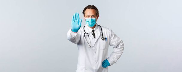 Covid-19, zapobieganie wirusom, pracownikom służby zdrowia i koncepcji szczepień. Poważny lekarz w masce medycznej i rękawicach wyciąga rękę, aby pokazać stop, zapobieganie, nie zgadzają się, lub ograniczyć, żądać społecznego dystansu - Zdjęcie, obraz