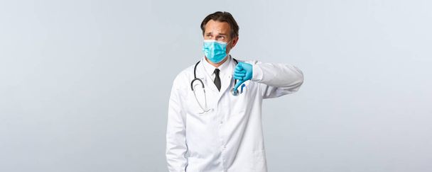 Covid-19, virüsü önleme, sağlık çalışanları ve aşı konsepti. Hüsrana uğramış tıbbi maskeli erkek doktor, eldivenler baş parmağını aşağı gösteriyor ve sol üst köşedeki tanıtıma bakıyor. - Fotoğraf, Görsel