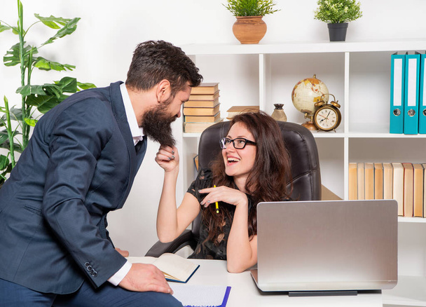 Smiling businesswoman pulling businessman sitting on office desk by beard, flirting - Fotoğraf, Görsel