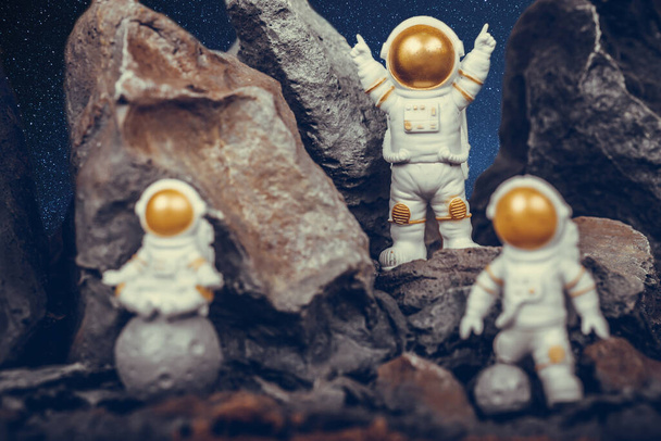 Altın vizörlü bir astronot ve uzay arkaplanlı kaya yüzeyinde beyaz bir uzay giysisi. Ay 'da bir astronot oturmuş meditasyon yaparken diğer astronotlar başarıyı kutluyor.. - Fotoğraf, Görsel