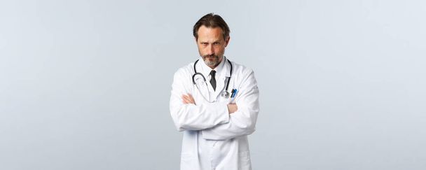 Covid-19, Coronavirus-Ausbruch, Gesundheitspersonal und Pandemiekonzept. Ernsthaft enttäuschter Arzt in weißem Mantel, Kreuzarmbrust und Blick unter die Stirn - Foto, Bild