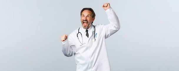 Covid-19, koronavírus járvány, egészségügyi dolgozók és világjárvány koncepció. Lelkes férfi orvos fehér köpenyben felemeli a kezét, és kiabál igen, néz sport mérkőzés, győzelem, diadal - Fotó, kép