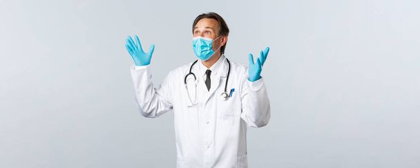 Covid-19, πρόληψη του ιού, εργαζόμενοι στον τομέα της υγείας και την έννοια του εμβολιασμού. Ενθουσιασμένος και ανακουφισμένος άνδρας γιατρός με ιατρική μάσκα, γάντια, σηκώνοντας τα χέρια ψηλά χαρούμενος, κοιτάζοντας πάνω αριστερή γωνία ευχαριστημένος - Φωτογραφία, εικόνα