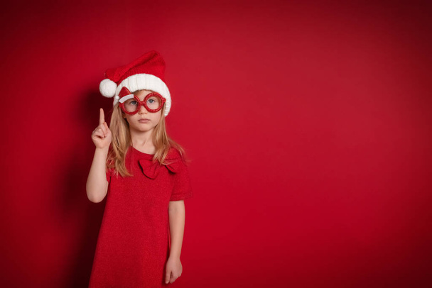 poważna dziewczyna w Boże Narodzenie ubrania podniósł palec w górę stoi na czerwonym tle - Zdjęcie, obraz
