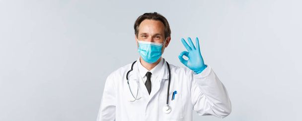 Covid-19 、ウイルス、医療、予防接種の概念を防止します。医療マスクと手袋の幸せな笑顔の医者は、すべての良いことを保証し、診療所でのサービスの安全性と品質を保証するとして大丈夫サインを示す - 写真・画像