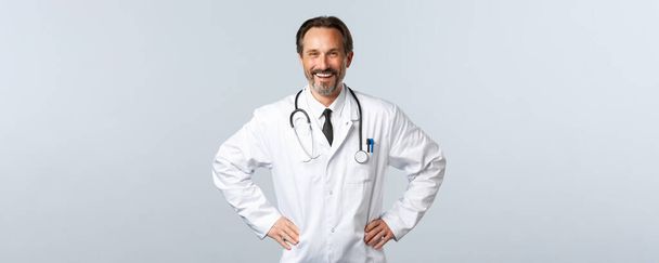 Covid-19, ξέσπασμα του ιού της στέψης, επαγγελματίες υγείας και πανδημία. Ενθουσιαστικός χαμογελαστός άντρας γιατρός που χαίρεται να βοηθάει τους ασθενείς. Γιατρός με λευκό παλτό χαρούμενος που εργάζεται σε κλινική ή νοσοκομείο - Φωτογραφία, εικόνα