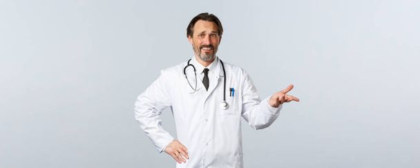Covid-19, Coronavirus-Ausbruch, Gesundheitspersonal und Pandemiekonzept. Skeptischer und verwirrter Arzt im weißen Kittel, der mit Bestürzung oder Ironie nach rechts zeigt, grimmig enttäuscht - Foto, Bild