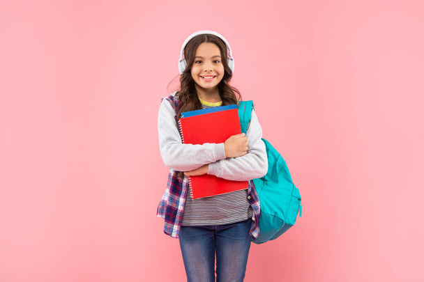 fröhliches Kind hört Musik in Kopfhörern mit Schultasche und Arbeitsheft auf rosa Hintergrund, eBook - Foto, Bild