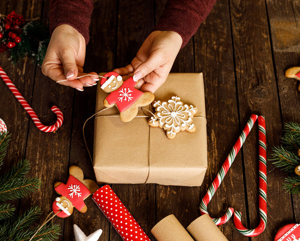Hands of a woman decorating a Christmas gift with snow flake shaped cookie and gingerbread Santa, winter holidays wooden background concept - 写真・画像
