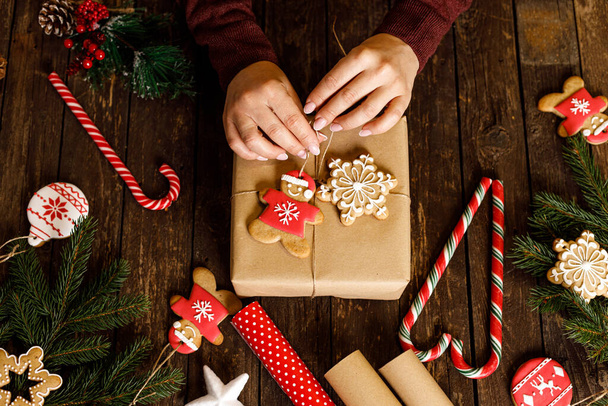Hands of a woman decorating a Christmas gift with snow flake shaped cookie and gingerbread Santa, winter holidays wooden background concept - Photo, image