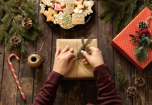 Hands of a woman decorating a Christmas gift with conifer branch on rustic wooden table full with cookies in festive winter holiday motives and shapes, merry Christmas concept - Foto, immagini
