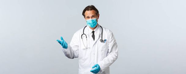Covid-19, Prävention von Viren, Gesundheitspersonal und Impfkonzept. Seriös aussehender Arzt in medizinischer Maske und Handschuhen unterhält sich mit Patienten, erklärt Maßnahmen während der Coronavirus-Pandemie - Foto, Bild