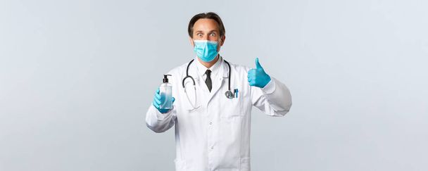 Covid-19, virüsü önleme, sağlık çalışanları ve aşı konsepti. Tıbbi maske ve eldiven takan heyecanlı doktor, yeni el dezenfektanı bulunca baş parmağını kaldır, hijyen ürünlerini tavsiye et. - Fotoğraf, Görsel