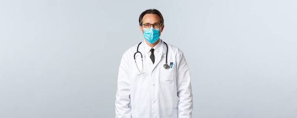Covid-19, zapobieganie wirusom, pracownikom służby zdrowia i koncepcji szczepień. Poważny pewny siebie lekarz w białym płaszczu i masce medycznej, noszący okulary, gotowy do leczenia pacjenta z grypą - Zdjęcie, obraz