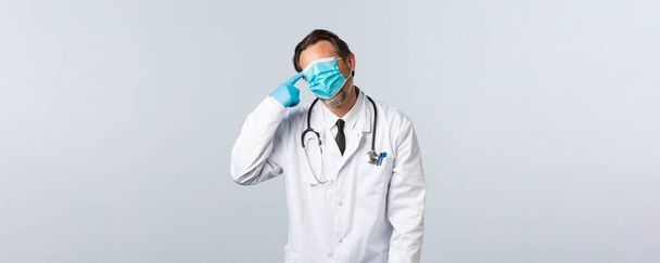 Covid-19, πρόληψη του ιού, εργαζόμενοι στον τομέα της υγείας και την έννοια του εμβολιασμού. Κουρασμένη και ταραγμένη γιατρός καταπολέμηση coronavirus, εξαντλημένη δουλειά, τραβήξτε μάσκα σε ολόκληρο το πρόσωπο και να πυροβολήσει χειρονομία όπλο κοντά στο κεφάλι - Φωτογραφία, εικόνα