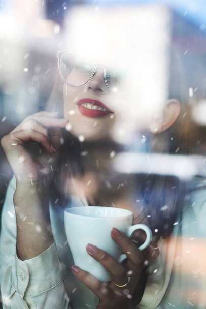 Impresionante mujer morena con lápiz labial rojo mantiene caliente con una taza de café caliente en un restaurante. Foto a través de la ventana en tiempo nevado - Foto, Imagen