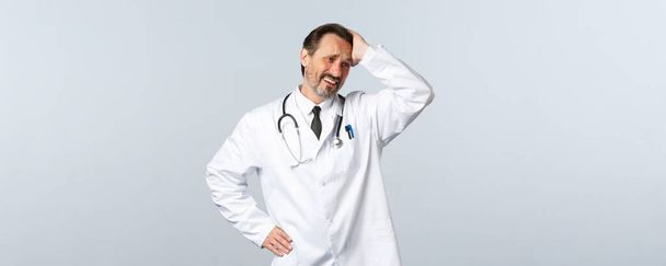 Covid-19, ξέσπασμα του ιού της στέψης, επαγγελματίες υγείας και πανδημία. Ταραγμένος και ανήσυχος απελπισμένος άντρας γιατρός με λευκό παλτό, αγγίζοντας το κεφάλι και γκρινιάζοντας, κοίτα αλλού θλιμμένος. - Φωτογραφία, εικόνα