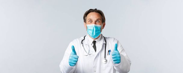 Covid-19, preventing virus, healthcare workers and vaccination concept. Excited and surprised happy doctor in medical mask and gloves, showing thumbs-up thrilled, hear great news, agree and approve - Photo, image