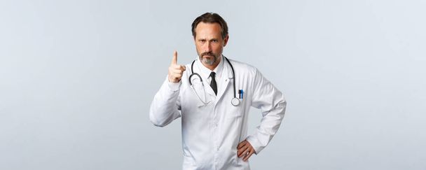 Covid-19, Coronavirus-Ausbruch, Gesundheitspersonal und Pandemiekonzept. Ernsthafter strenger Arzt runzelt die Stirn, schaut enttäuscht und besorgt, schüttelt den Finger, um Patienten schlechtes Benehmen vorzuwerfen - Foto, Bild