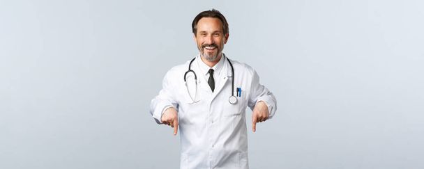 Covid-19, Coronavirus-Ausbruch, Gesundheitspersonal und Pandemiekonzept. Fröhlich lächelnder Arzt im weißen Kittel, der zum Test in die Klinik einlädt, Zeigefinger nach unten, Werbung - Foto, Bild