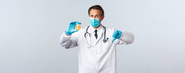 Covid-19, virüsü önleme, sağlık çalışanları ve aşı konsepti. Tıbbi maske ve eldiven giymiş hayal kırıklığına uğramış bir doktor idrar örneği gösteriyor, baş parmağının düşmesi kötü bir test sonucu, beyaz bir geçmiş. - Fotoğraf, Görsel