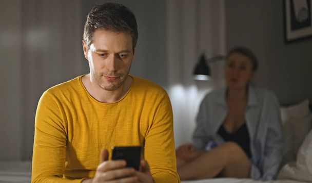 Telefoonverslaving concept. Man met smartphone, vrouw van streek op de achtergrond. - Foto, afbeelding