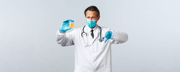 Covid-19, Prävention von Viren, Gesundheitspersonal und Impfkonzept. Enttäuschter Arzt in medizinischer Maske und Handschuhen zeigt Urinprobe, Daumen nach unten haben schlechtes Testergebnis, weißer Hintergrund - Foto, Bild