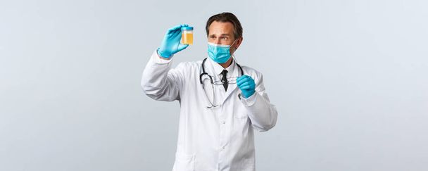 Covid-19, virüsü önleme, sağlık çalışanları ve aşı konsepti. Tıbbi maskeli ve eldivenli ciddi görünümlü bir doktor idrar örneğini inceler, düşünceli ve konsantre görünür. - Fotoğraf, Görsel