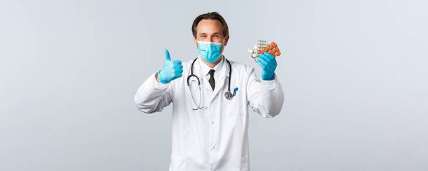 Covid-19, zapobieganie wirusom, pracownikom służby zdrowia i koncepcji szczepień. Zadowolony przystojny lekarz uśmiechnięty, nosić maskę medyczną i rękawiczki, kciuk do góry jak polecam pigułki, pokazać dobre leki - Zdjęcie, obraz