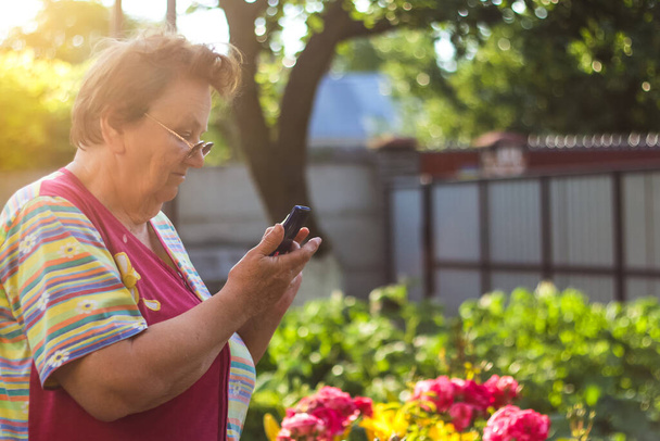 Een 65-jarige vrouw kijkt naar de telefoon in de tuin Oma leert hoe de telefoon te gebruiken.Portret van goed uitziende volwassen vrouw met behulp van smart phone typen berichten via sociaal netwerk. Zachte focus. - Foto, afbeelding