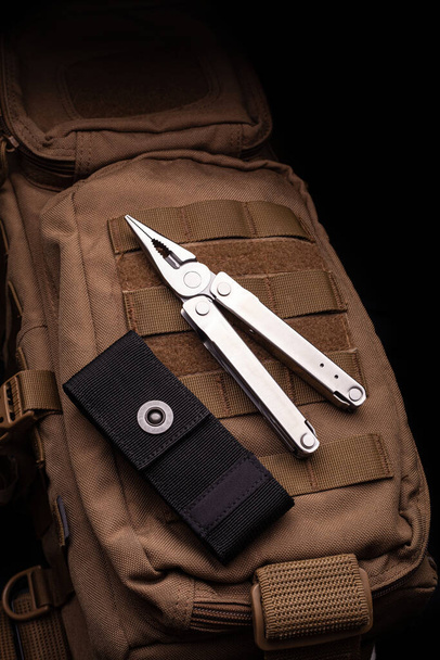 Modern multitool with many tools. A portable multitasking tool lie on a desert-colored military backpack. - 写真・画像