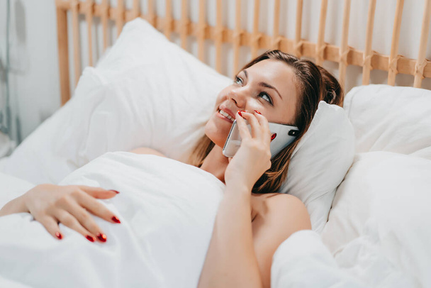 Woman talking on smartphone while lying in comfortable bed. Lady holding mobile in light cozy bedroom. Addiction to technology, morning rituals concept. - Photo, Image