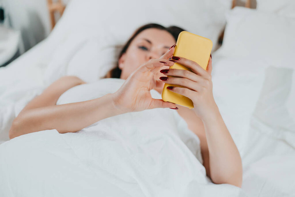 Woman holds smartphone in hands while lying in comfortable bed. Lady with mobile in light cozy bedroom. Addiction to technology and social media, morning rituals concept. - Photo, Image